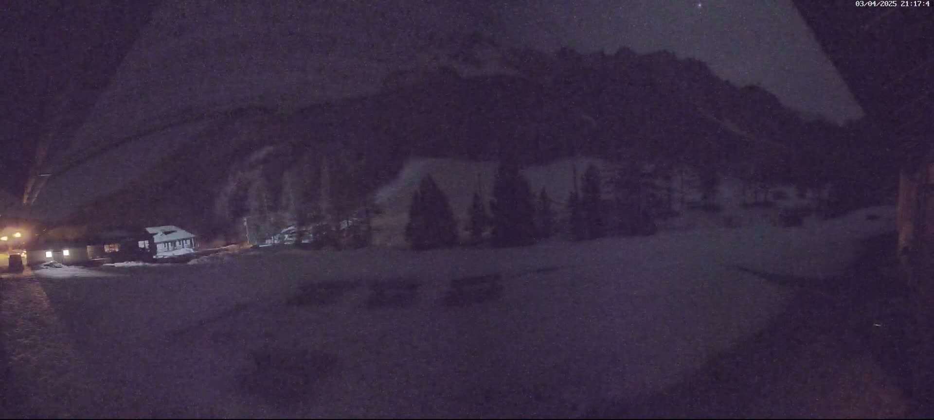 A nighttime, snowy scene shows a cluster of buildings nestled below a dark mountain range under a slightly hazy sky.