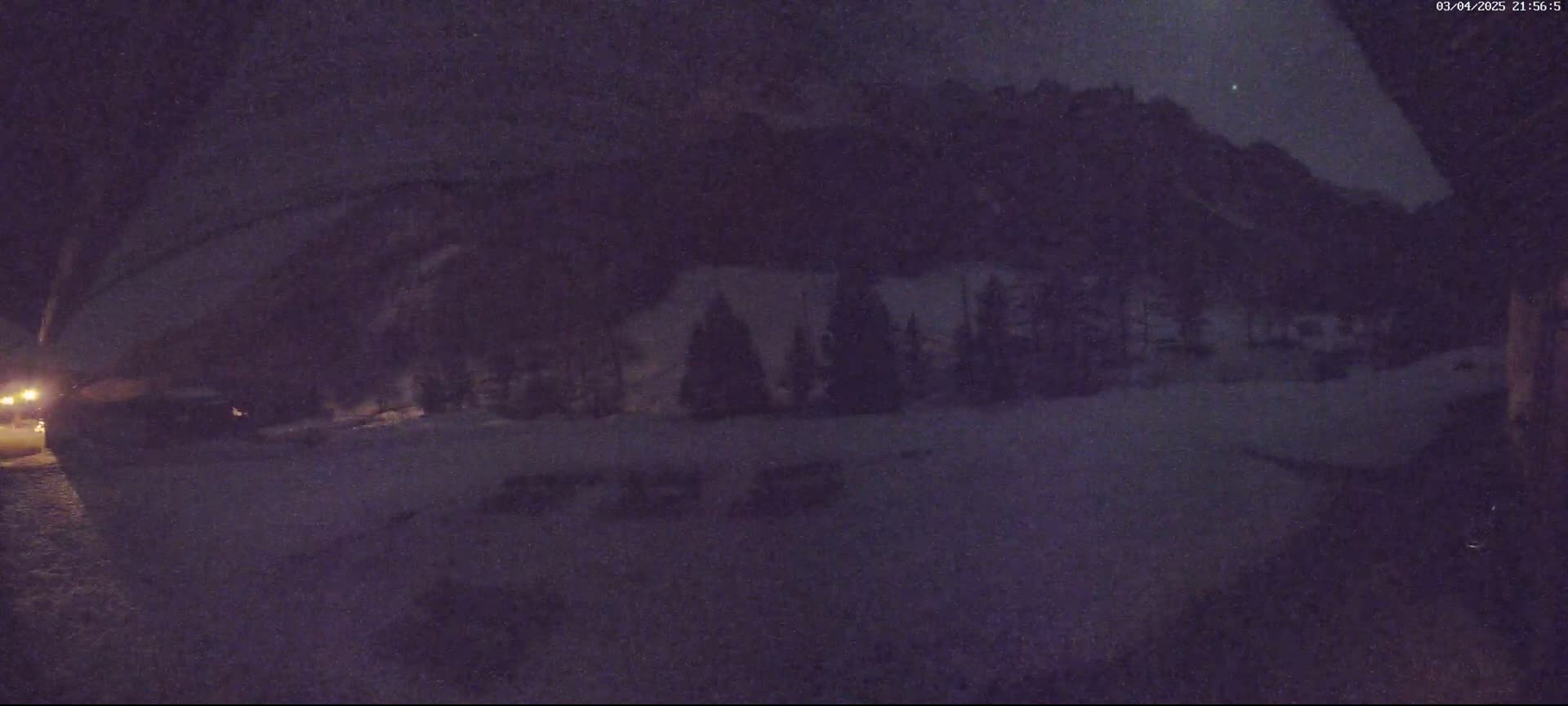 A nighttime, snowy scene shows dark, snow-covered ground in the foreground, a line of silhouetted trees in the middle ground, and a mountainside under a mostly clear night sky in the background.