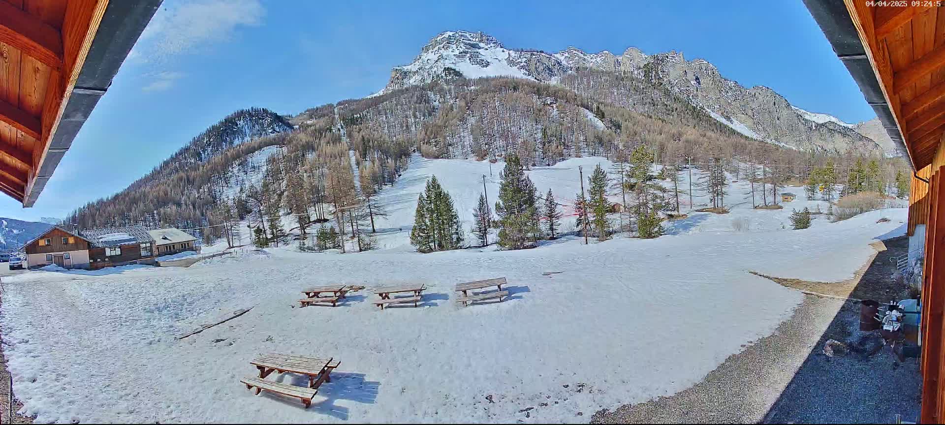 A sunny, snow-covered mountain landscape is visible from a wooden structure, featuring several picnic tables in the foreground and a small building in the mid-ground.