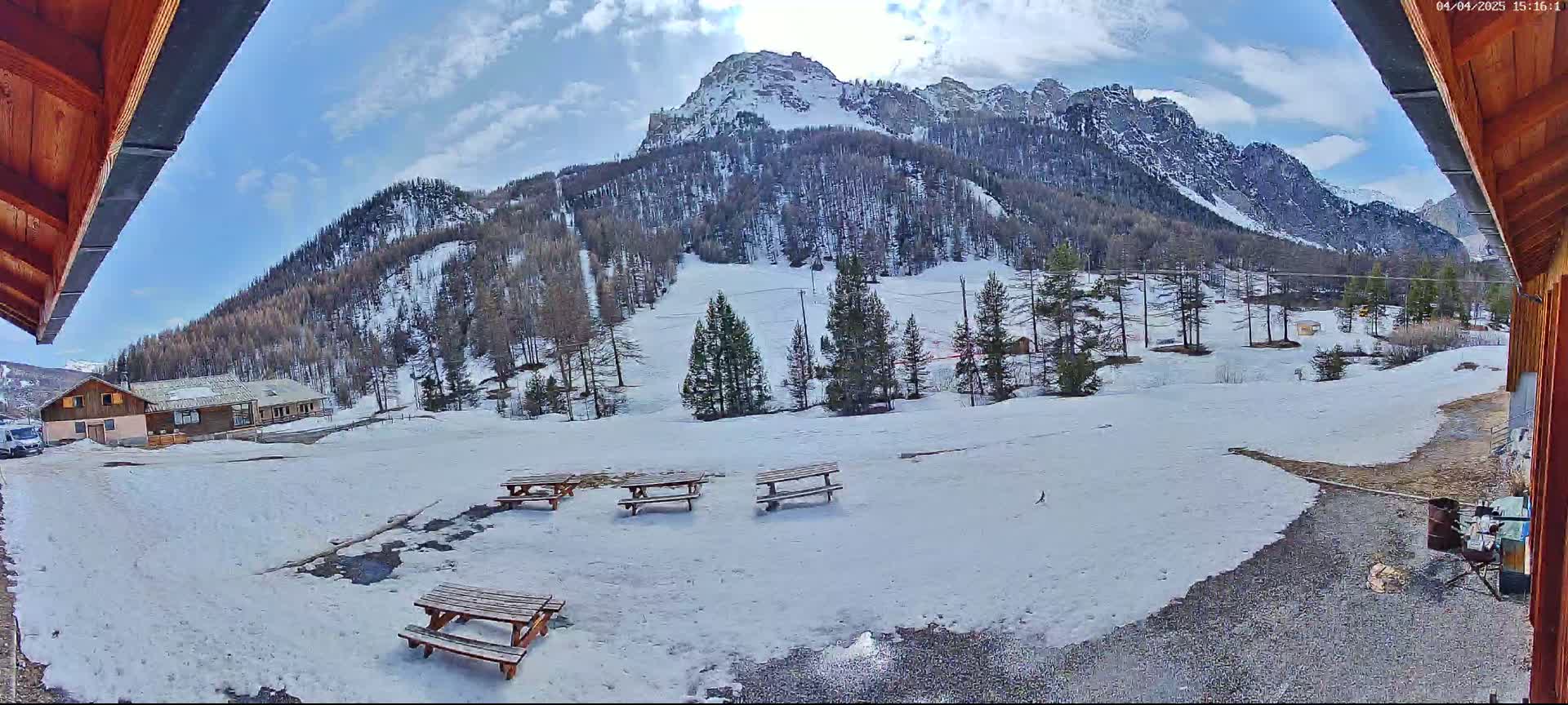 A snow-covered mountainside with several picnic tables in the foreground, viewed from under a wooden overhang on a partly cloudy day.