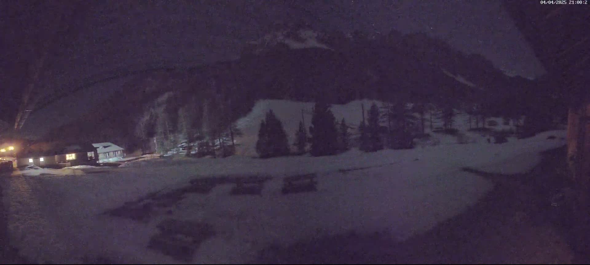 A snow-covered landscape at night shows a small building illuminated near a stand of dark evergreens beneath a mountain range.