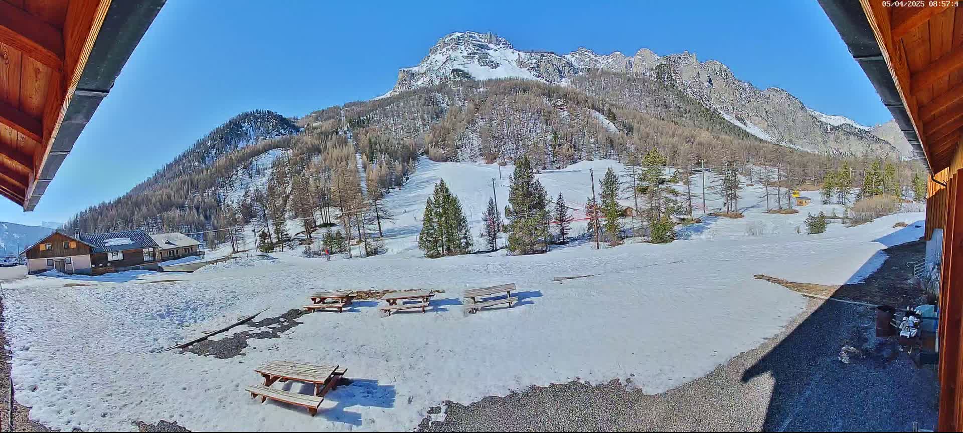 A sunny view from a wooden structure shows a snow-covered landscape with picnic tables, small buildings, and a mountainous backdrop.