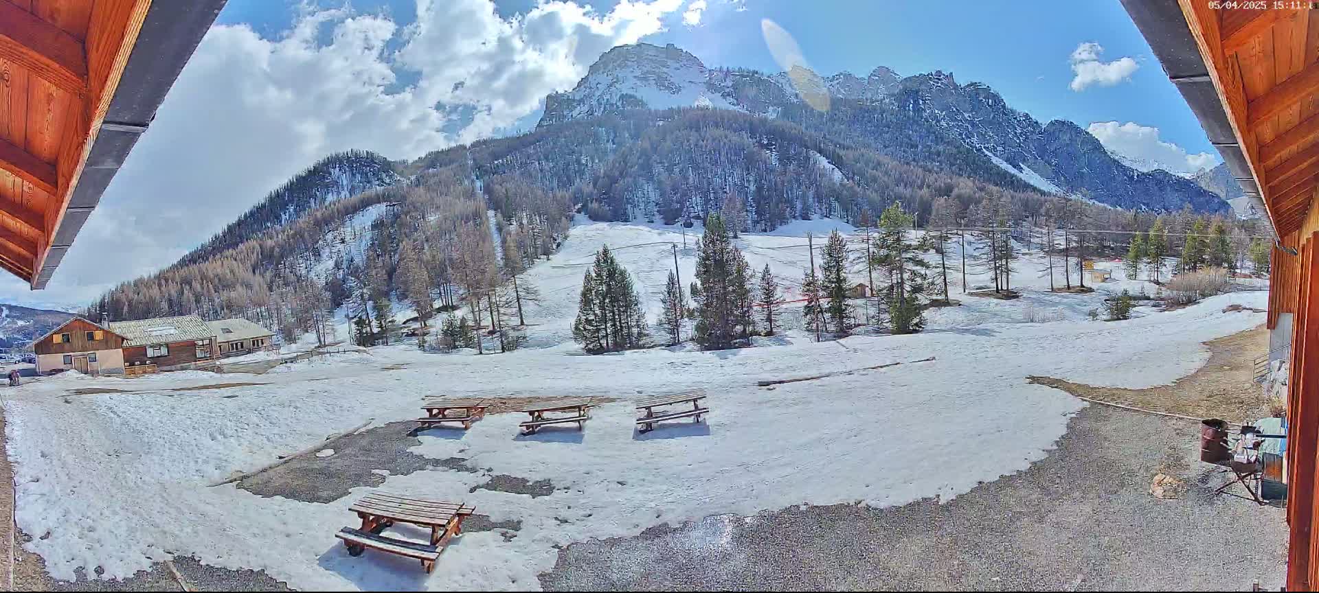 A snow-covered mountainside with several picnic tables sits in the foreground, viewed from under a wooden overhang on a partly sunny day.