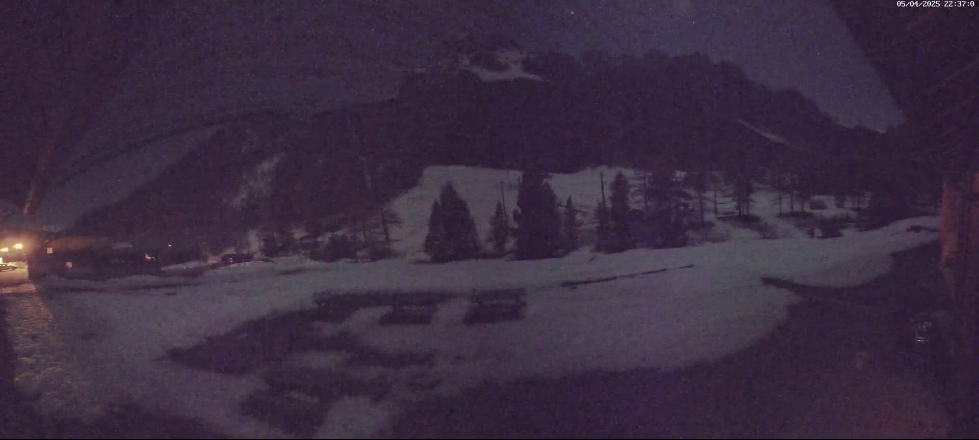 A nighttime snow-covered landscape shows a snow-covered parking lot, a few trees, and dark mountains in the background under a lightly snowing sky.
