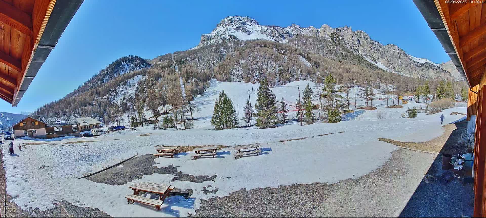 A sunny, snow-covered mountainside with several picnic tables in a gravel area, viewed from under a wooden roof.