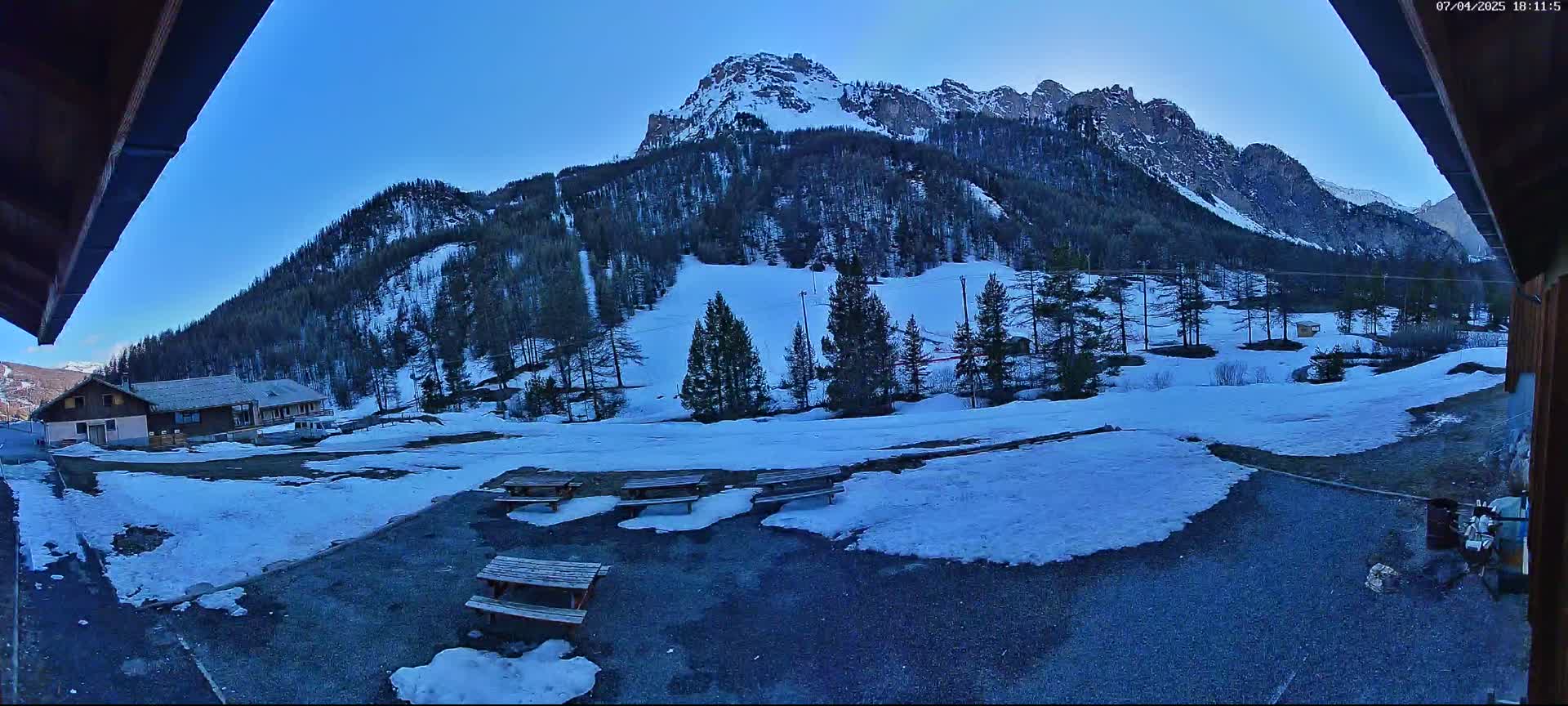A snow-covered mountainside with several picnic tables in the foreground under a clear blue sky.