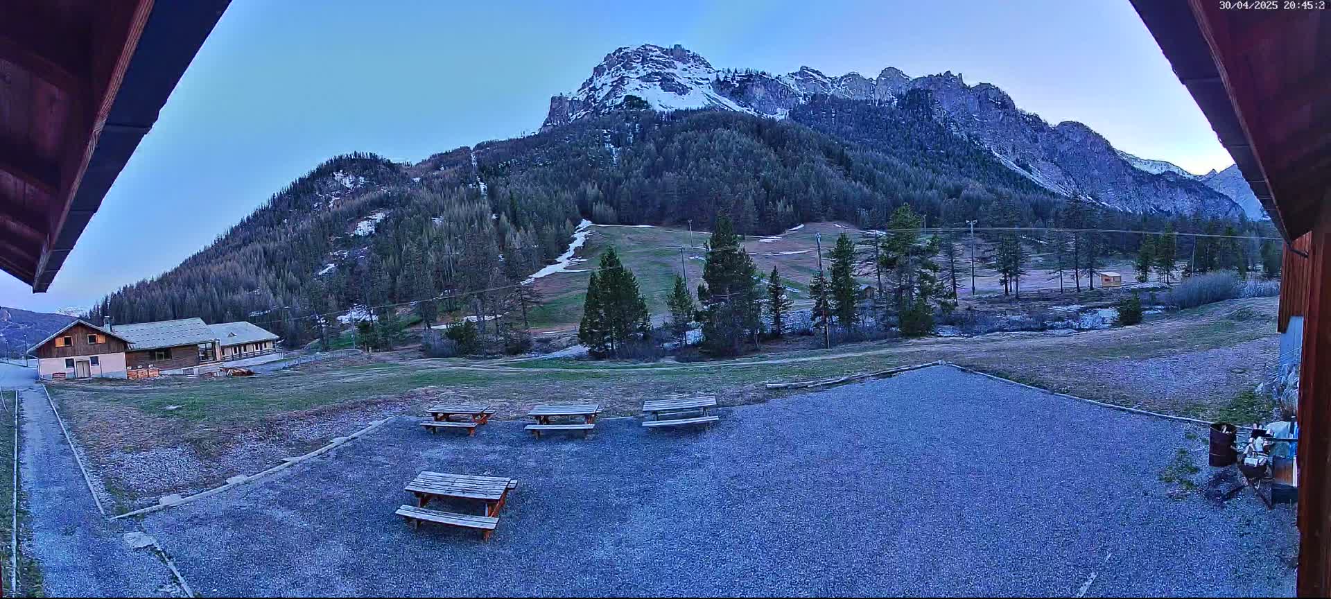 A gravel area with several picnic tables sits in front of a building, overlooking a grassy field and snow-capped mountains under a clear, twilight sky.