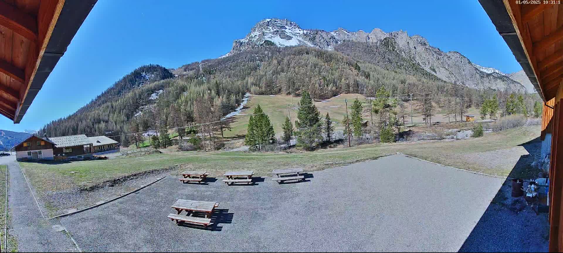 A gravel area with several picnic tables overlooks a grassy field, a small building, and a mountain range under a clear blue sky.