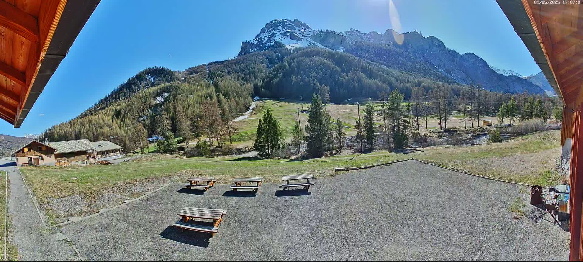 A sunny view from a wooden structure shows several picnic tables in a gravel area overlooking a grassy field, a building, and a snow-capped mountain range under a clear blue sky.