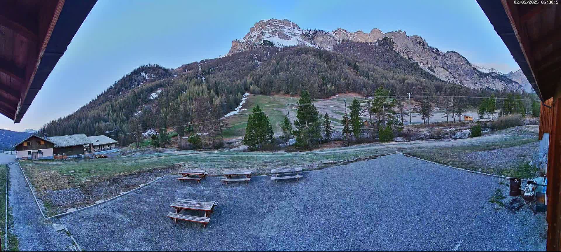 A gravel area with several picnic tables sits in front of a building, overlooking a grassy field, a sparsely wooded hillside with a ski slope, and a snow-capped mountain range under a clear, early morning sky.
