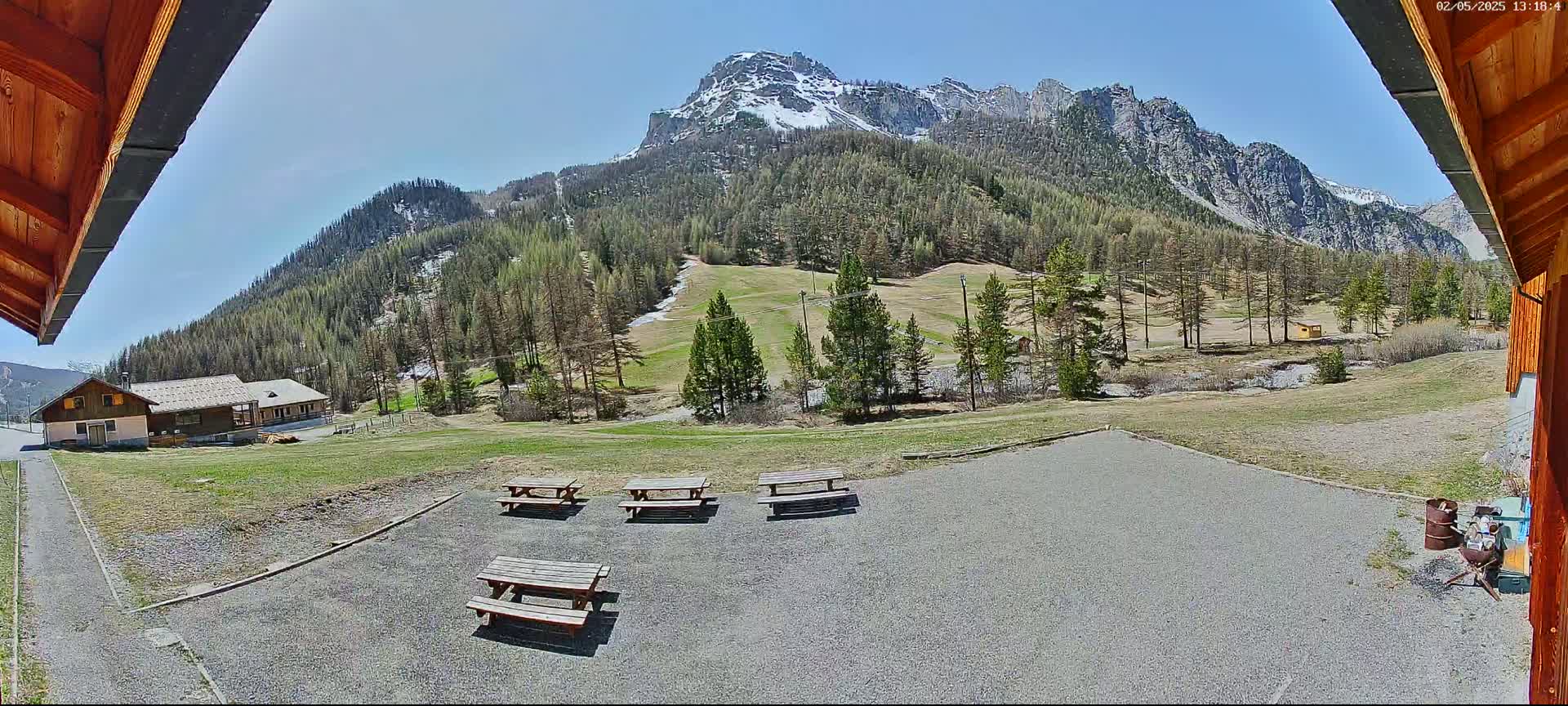 A sunny view of a grassy area with picnic tables, a small building, and snow-capped mountains in the background.