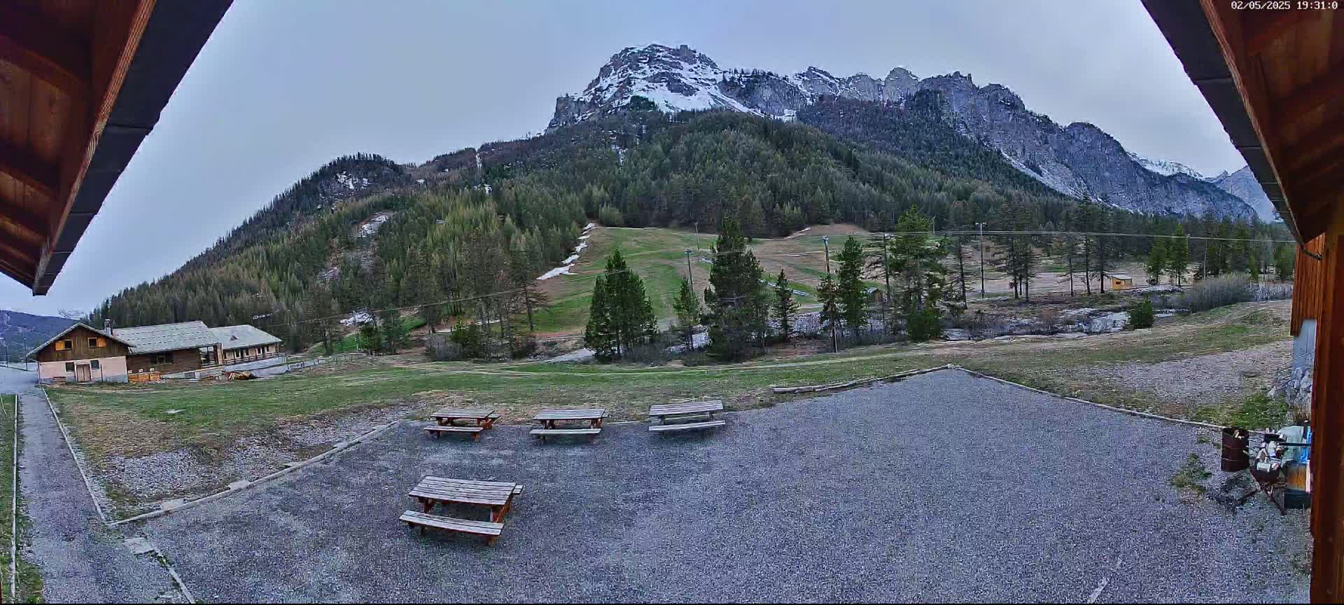 A gravel area with picnic tables sits before a small building and a grassy field, all overlooked by a snow-dusted mountain range under a mostly clear sky.