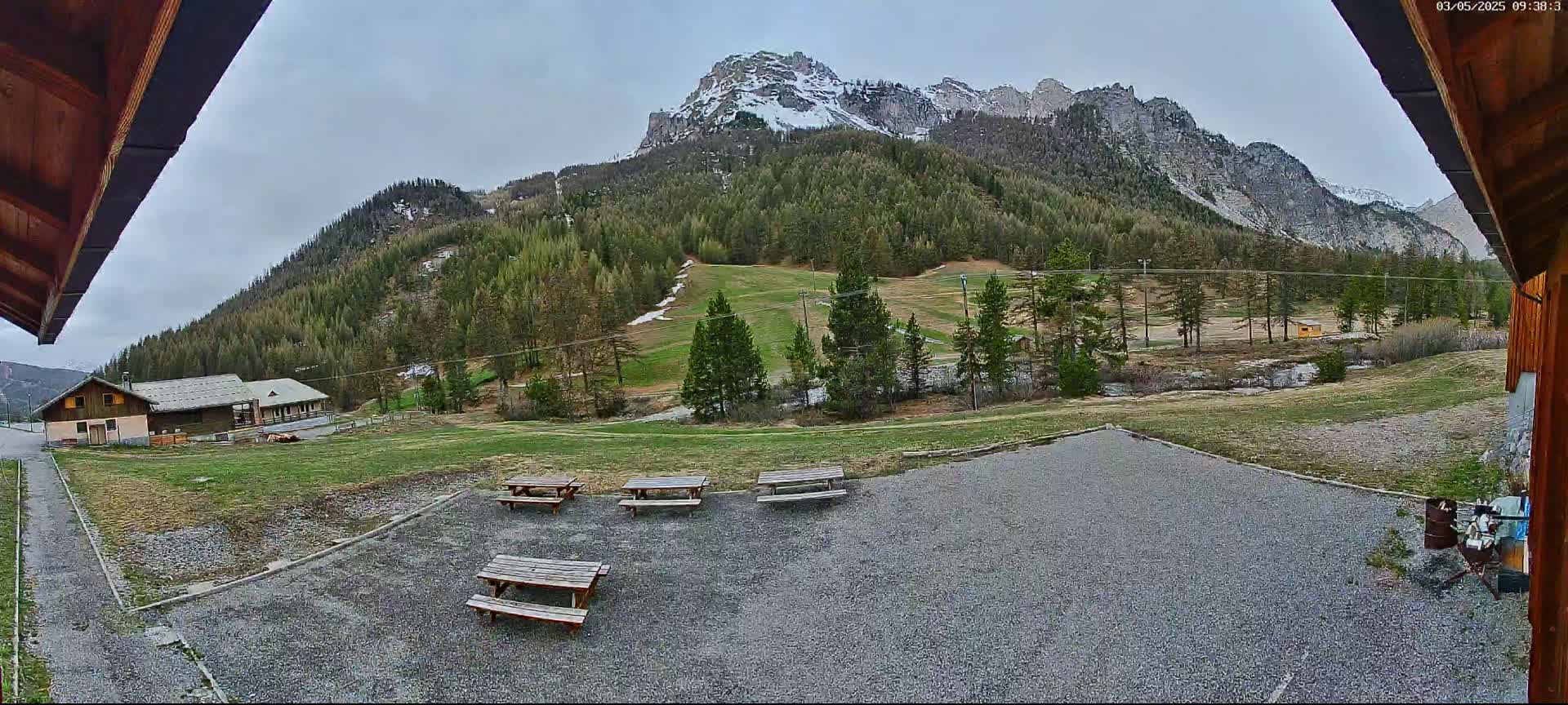 A gravel area with several picnic tables overlooks a grassy field, a small building, and a mountain range partially covered in snow under an overcast sky.