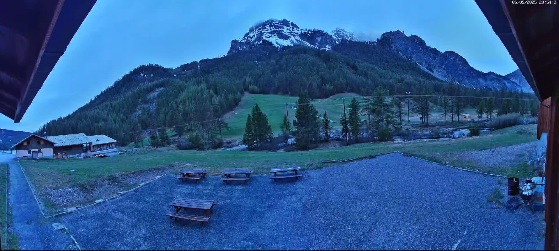 Several picnic tables sit in a gravel area overlooking a grassy field, a small building, and a snow-capped mountain under a twilight sky.
