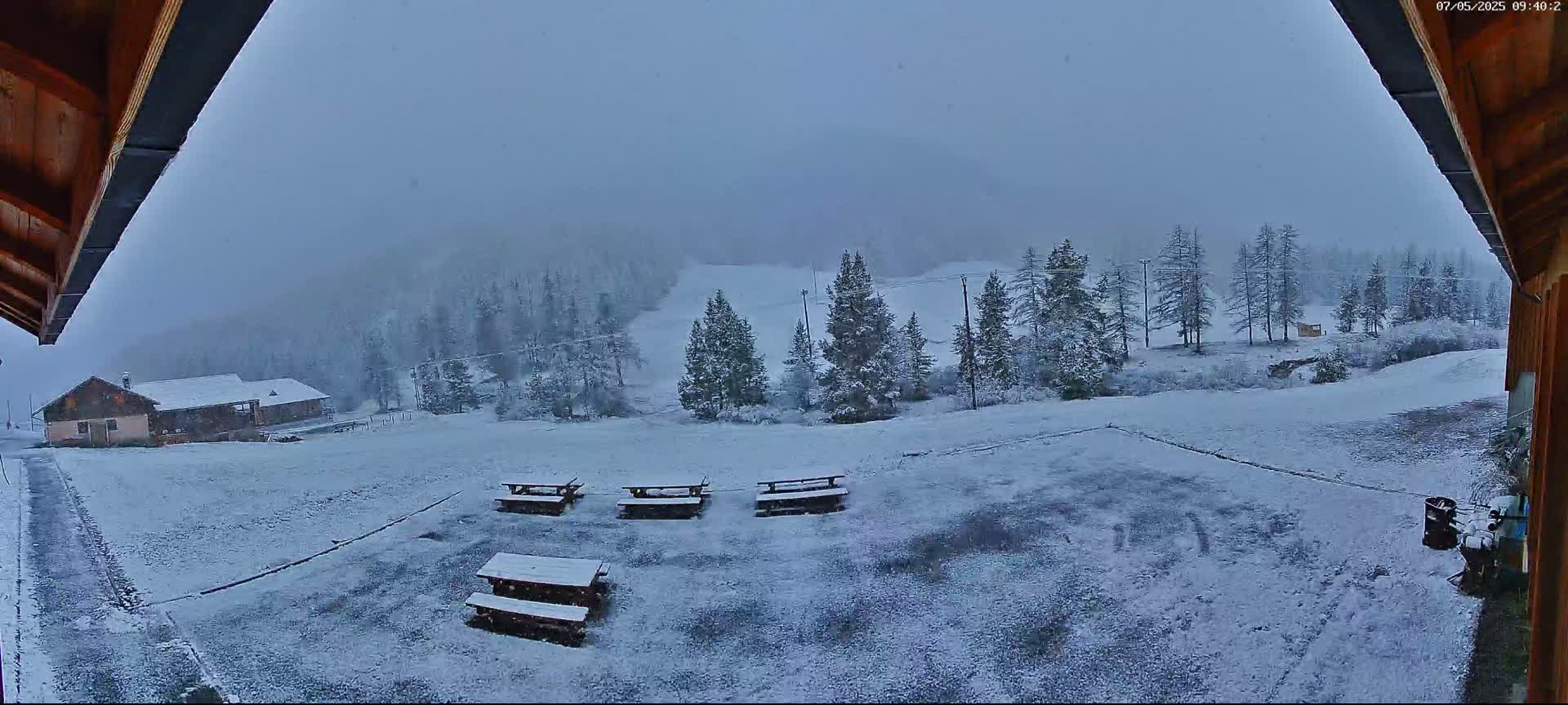 A snow-covered landscape with several picnic tables and a building in the distance is visible under a lightly snowing sky.