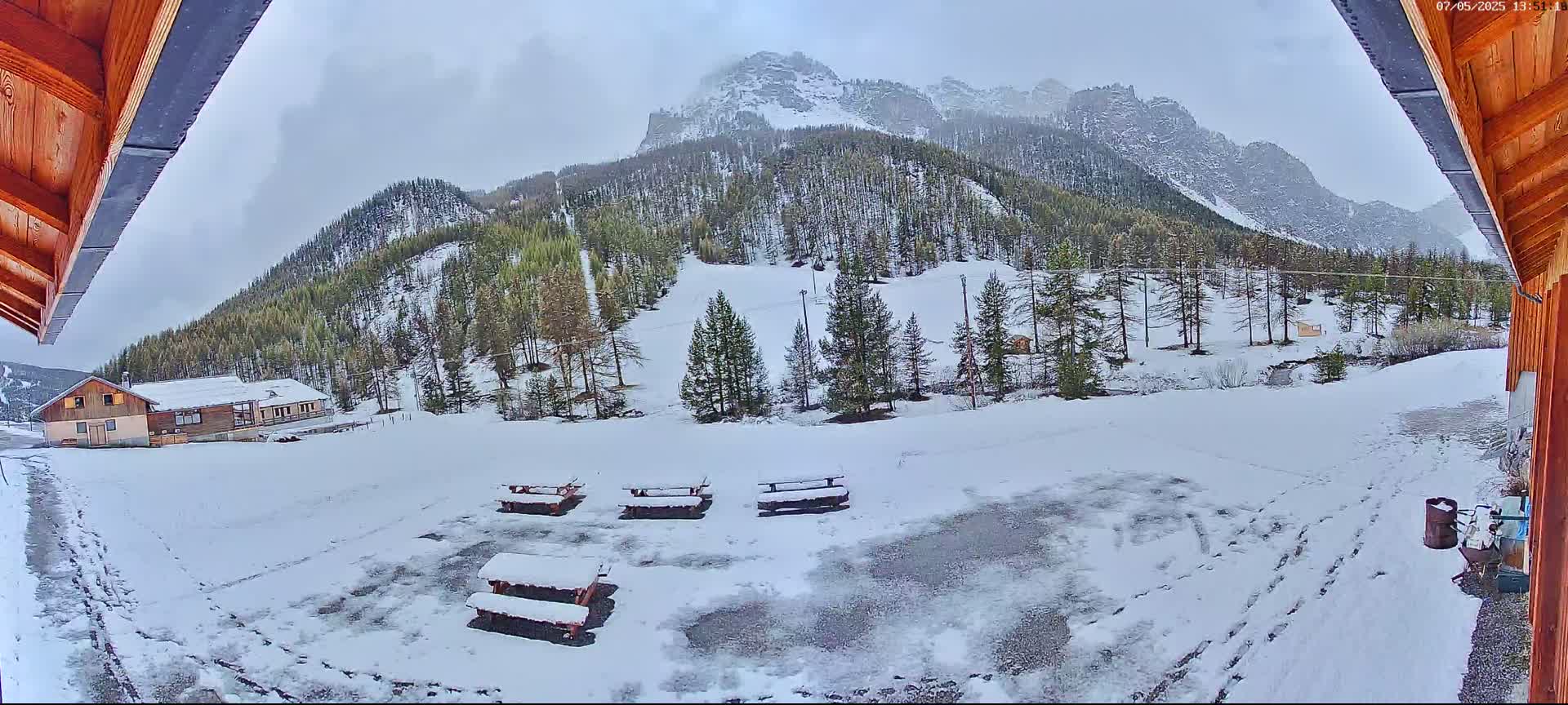A snow-covered landscape with several picnic tables, a building, and a mountain range in the background under an overcast sky.