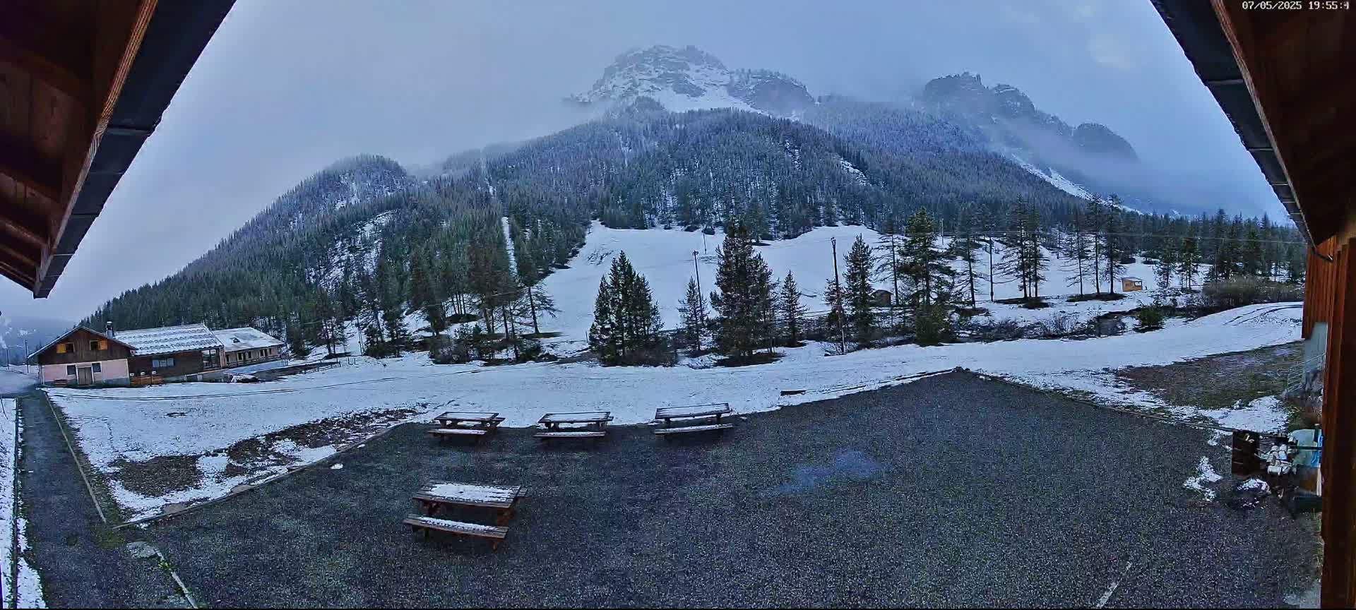 A snow-covered mountain landscape is viewed from a covered patio area with several picnic tables, under an overcast sky.