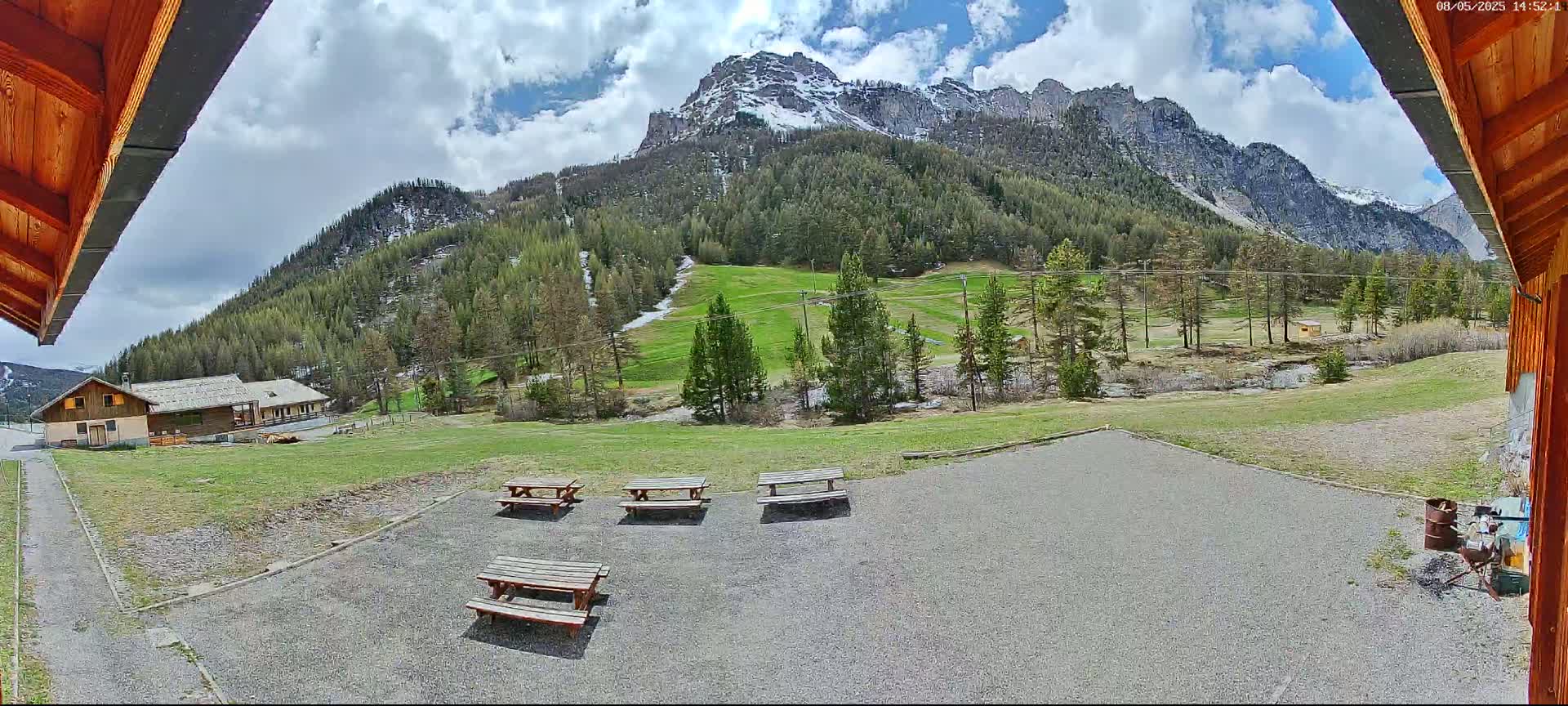 A partly cloudy day reveals a mountain with some snow near the peak, green hillsides dotted with trees and several picnic tables in a gravel area near a small building.