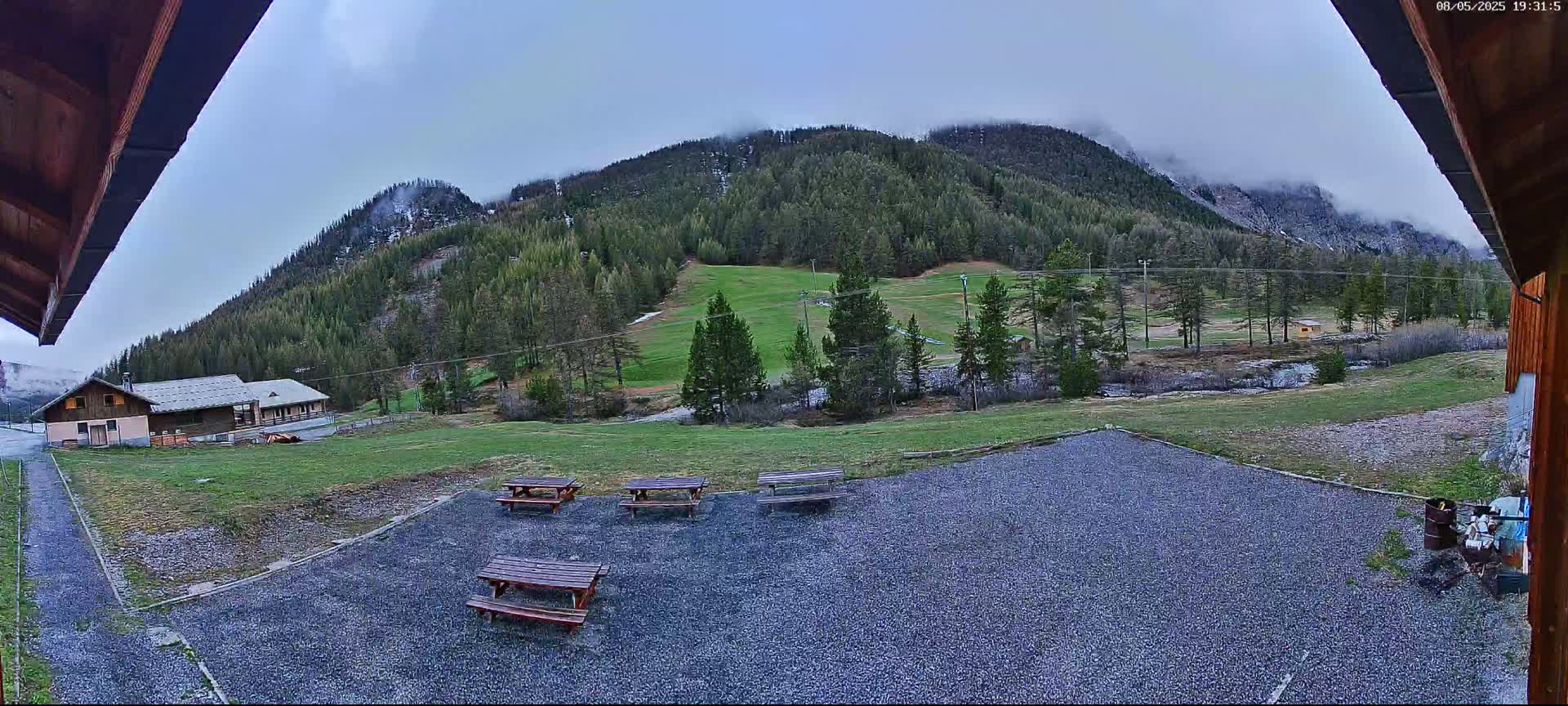 A gravel area with picnic tables overlooks a grassy field, a small building, and a mountain covered in patchy clouds and some snow.