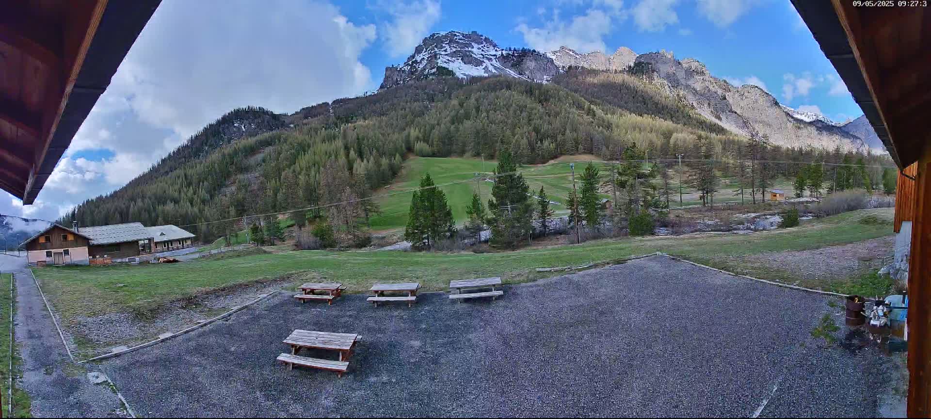 A gravel area with several picnic tables overlooks a grassy field, a building, and a mountain range with some snow under a partly cloudy sky.