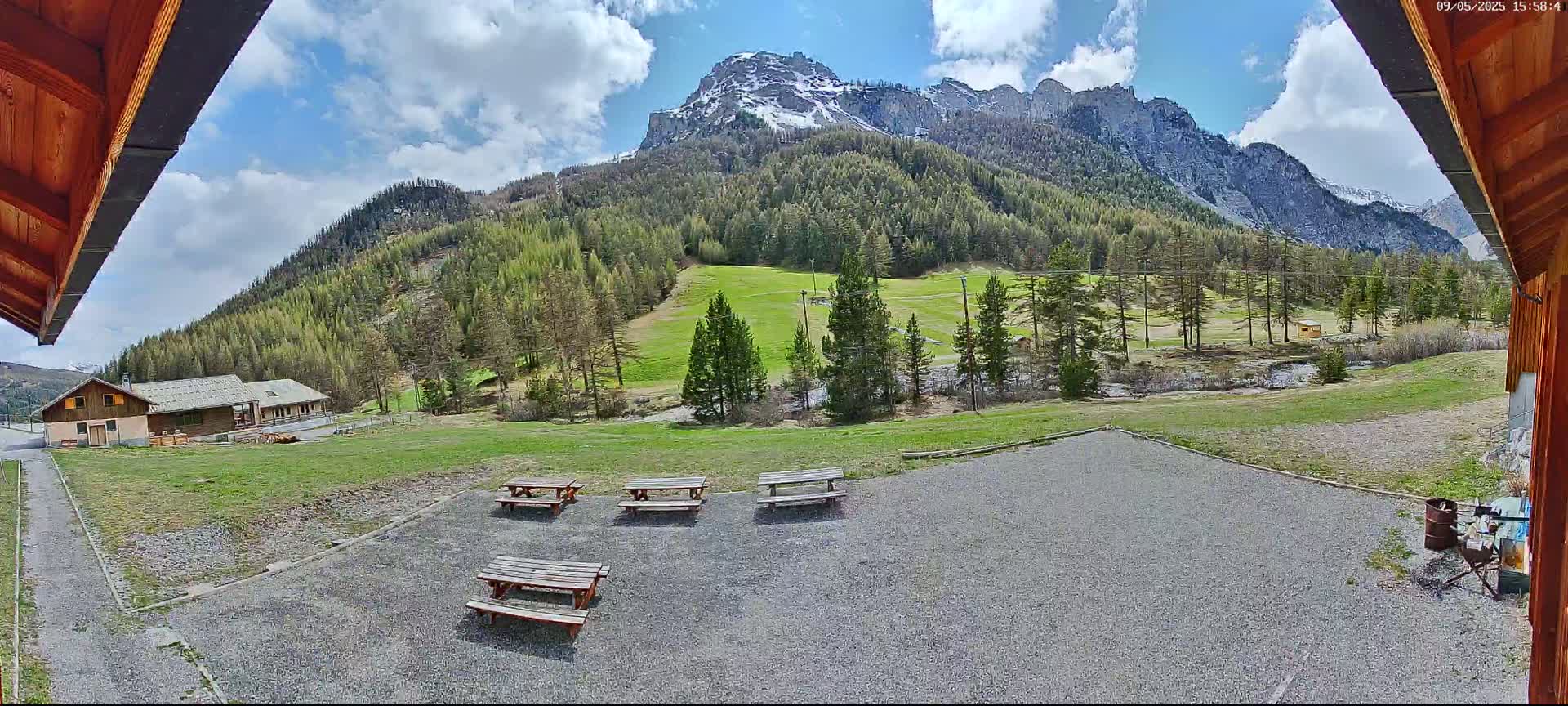 A gravel area with several picnic tables overlooks a grassy field, a small building, and a mountain range with some snow under a partly cloudy sky.