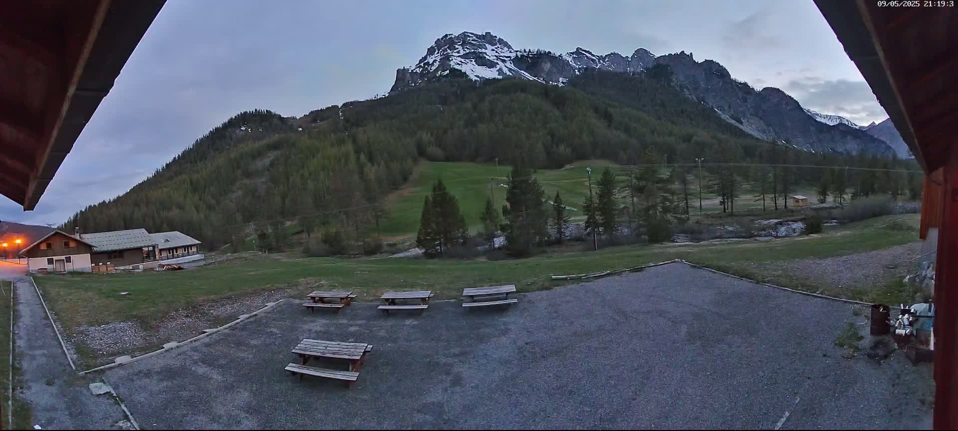 A gravel area with picnic tables sits before a grassy field and a mountain range partially covered in snow under a twilight sky.