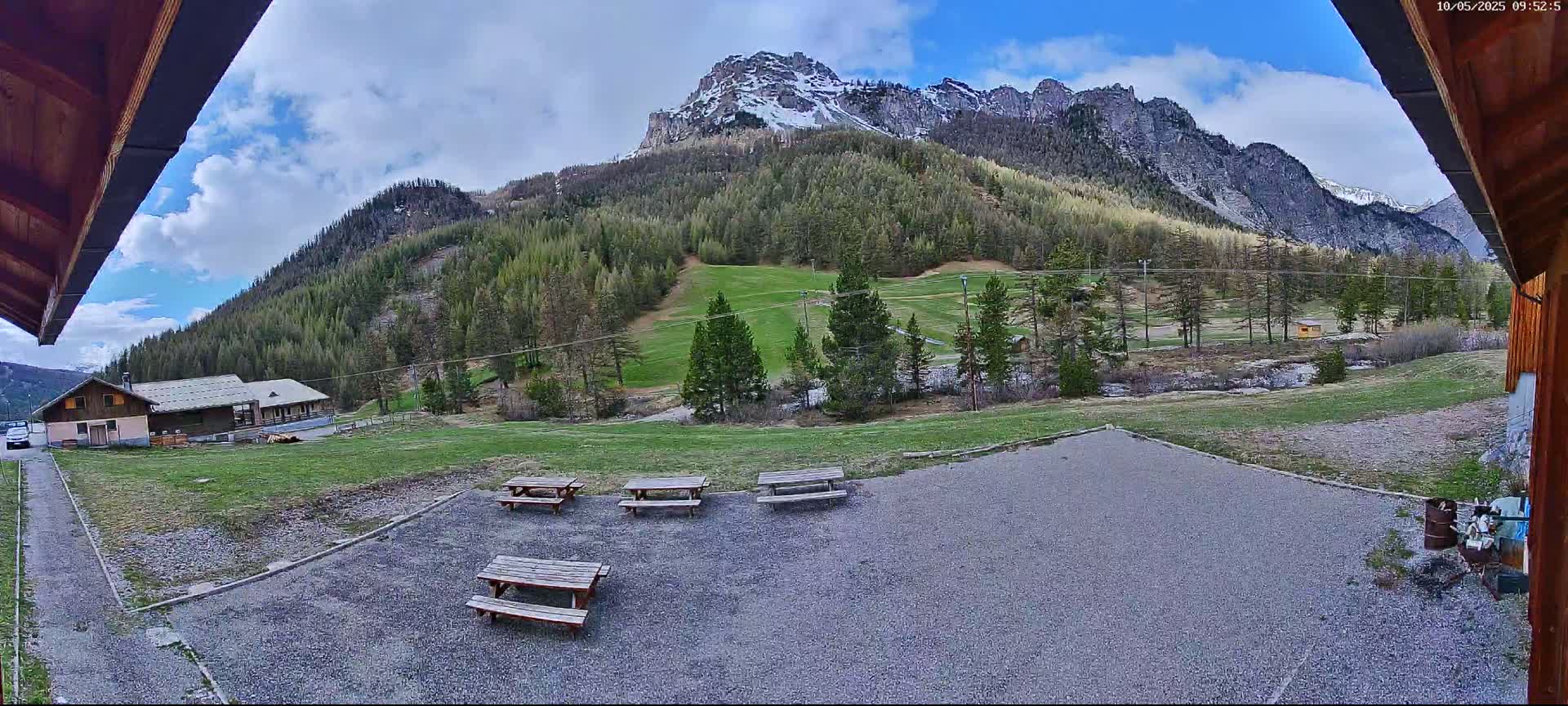 A partly cloudy day reveals a mountainside covered in evergreen trees, a grassy area with picnic tables, and a small building in the foreground, all viewed from under an overhang.
