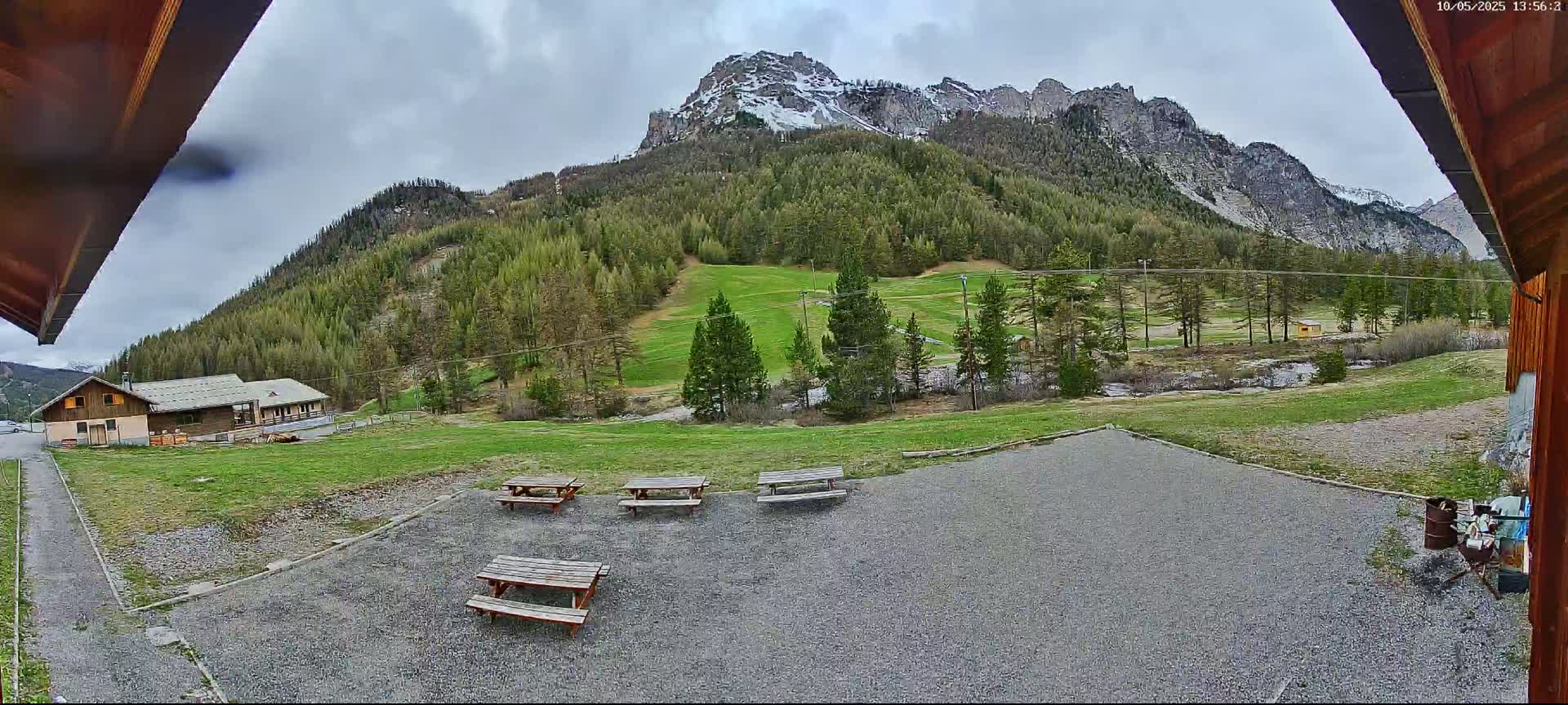 A gravel area with several picnic tables overlooks a grassy field, a small building, and a mountain range with patches of snow under an overcast sky.