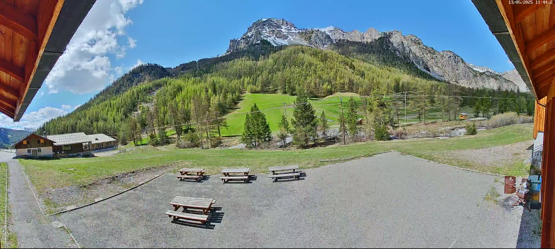 A gravel area with several picnic tables sits in front of a building, overlooking a grassy field, a forest of bright green trees, and a mountain range under a mostly sunny sky.