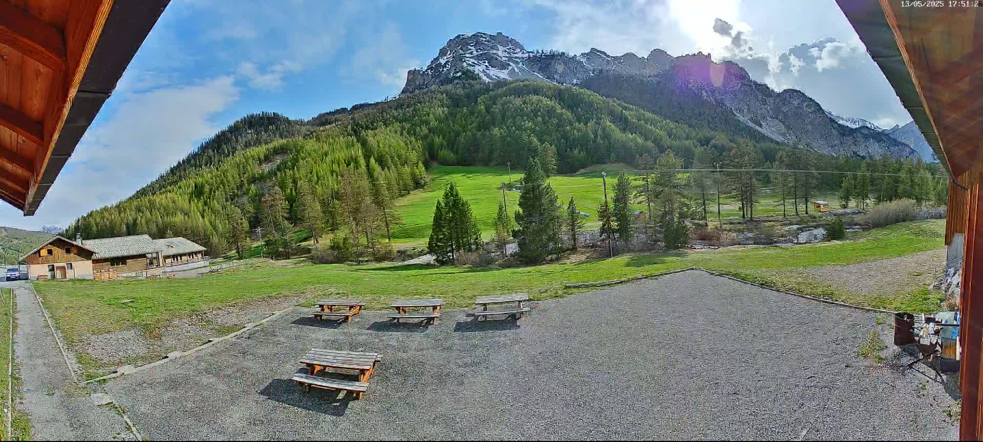 A gravel area with picnic tables sits in front of a building, overlooking a grassy field, a forest of pine trees, and snow-capped mountains under a partly sunny sky.