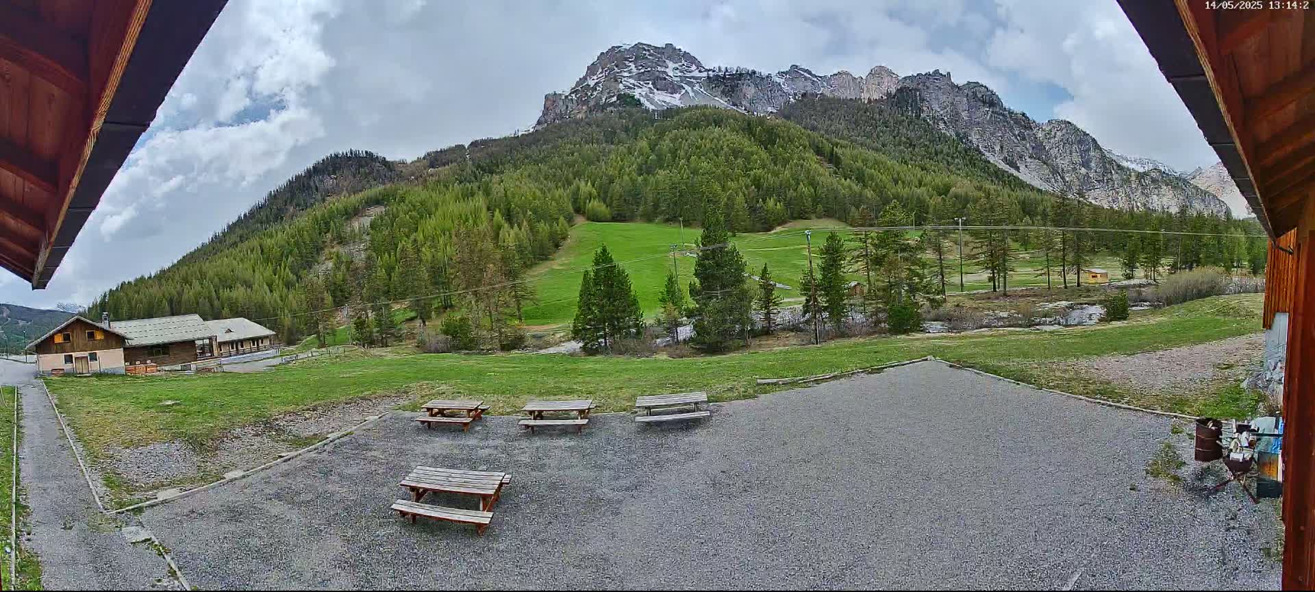 A gravel area with picnic tables overlooks a grassy field, a small building, and a mountain range under a partly cloudy sky.