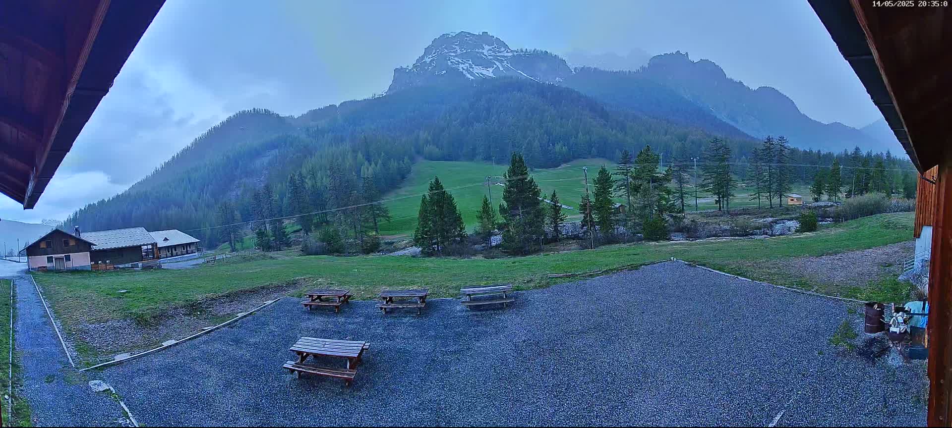 A gravel area with picnic tables sits in front of a building, overlooking a green valley and snow-capped mountains under a cloudy sky.