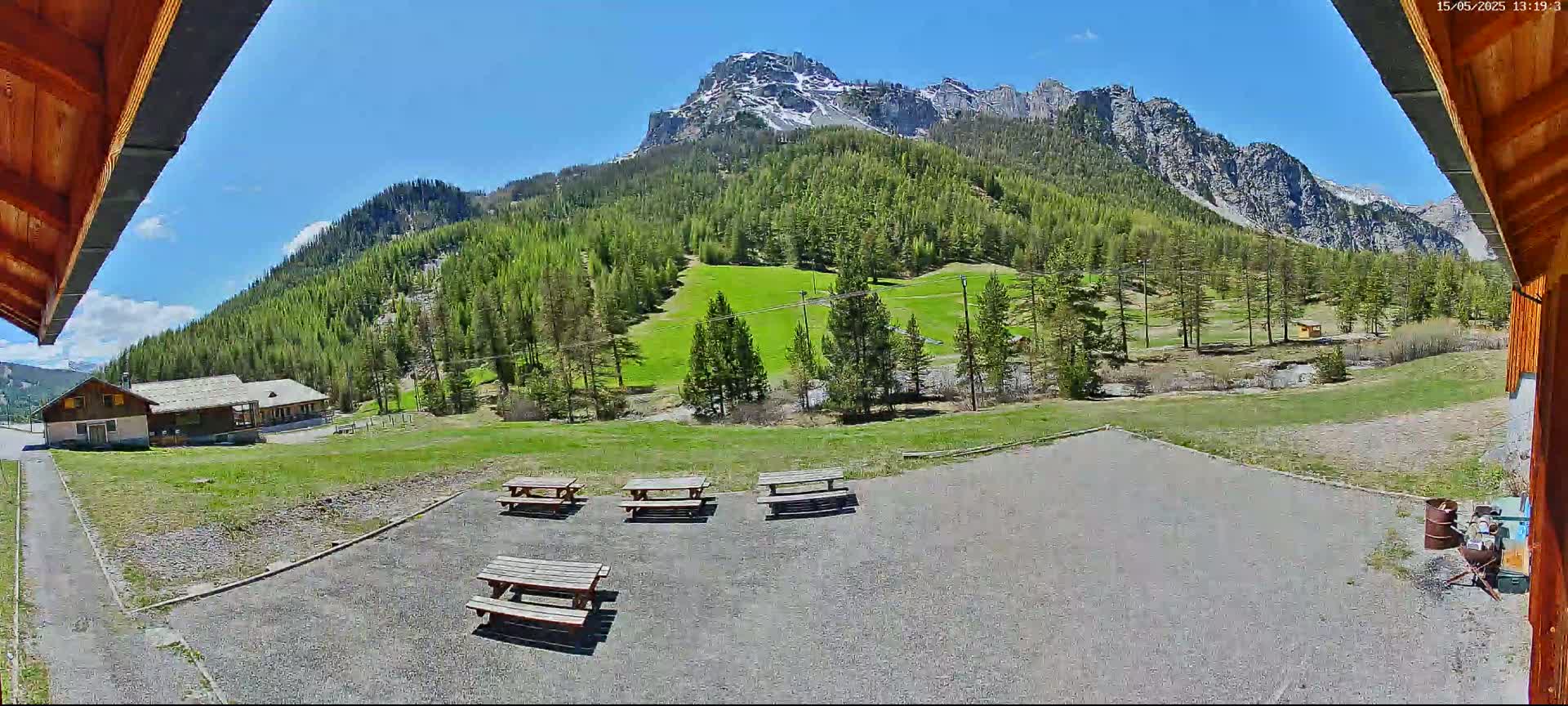 Several picnic tables sit in a gravel area overlooking a grassy valley dotted with evergreen trees and a snow-capped mountain range under a bright, sunny sky.
