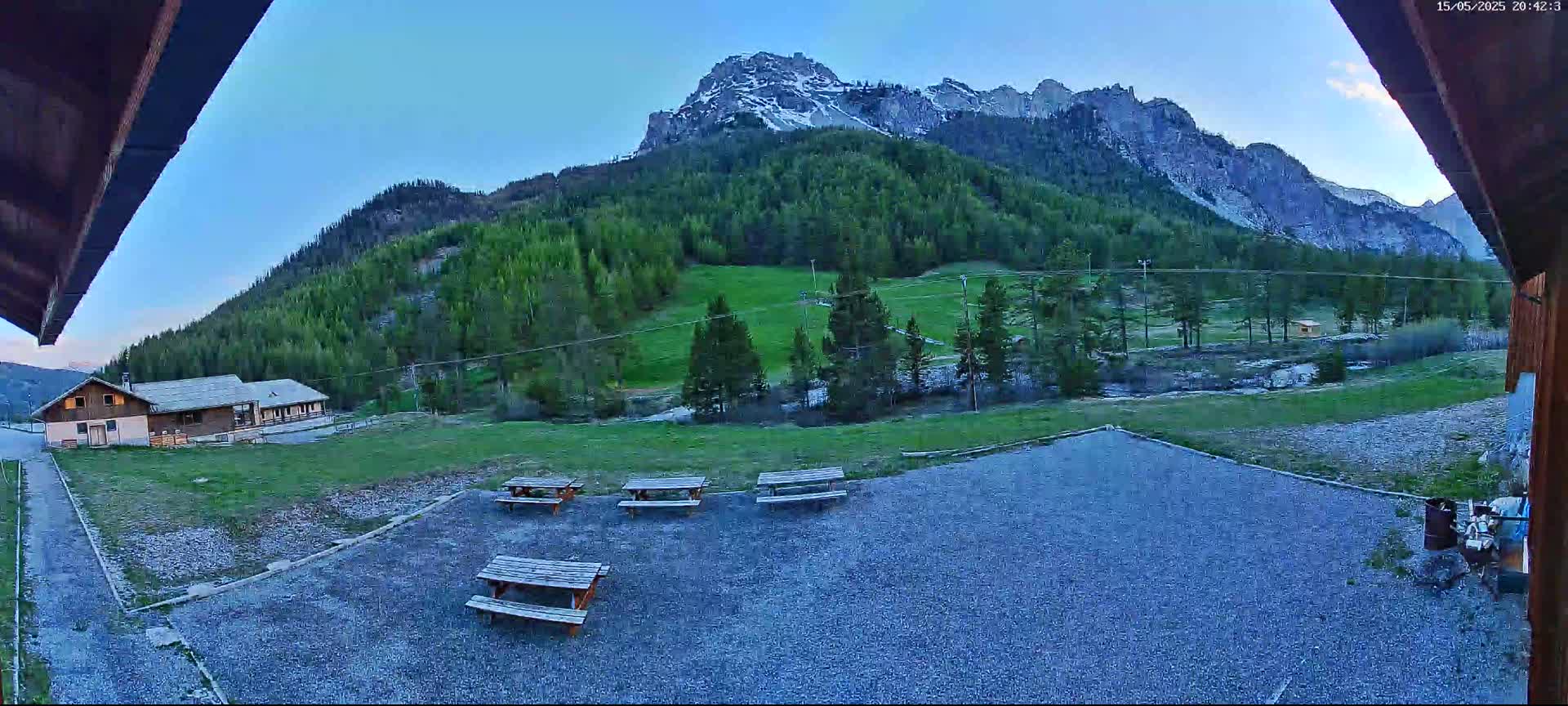 Several picnic tables sit in a gravel area overlooking a grassy field, a building, and a mountain range with some snow under a clear twilight sky.