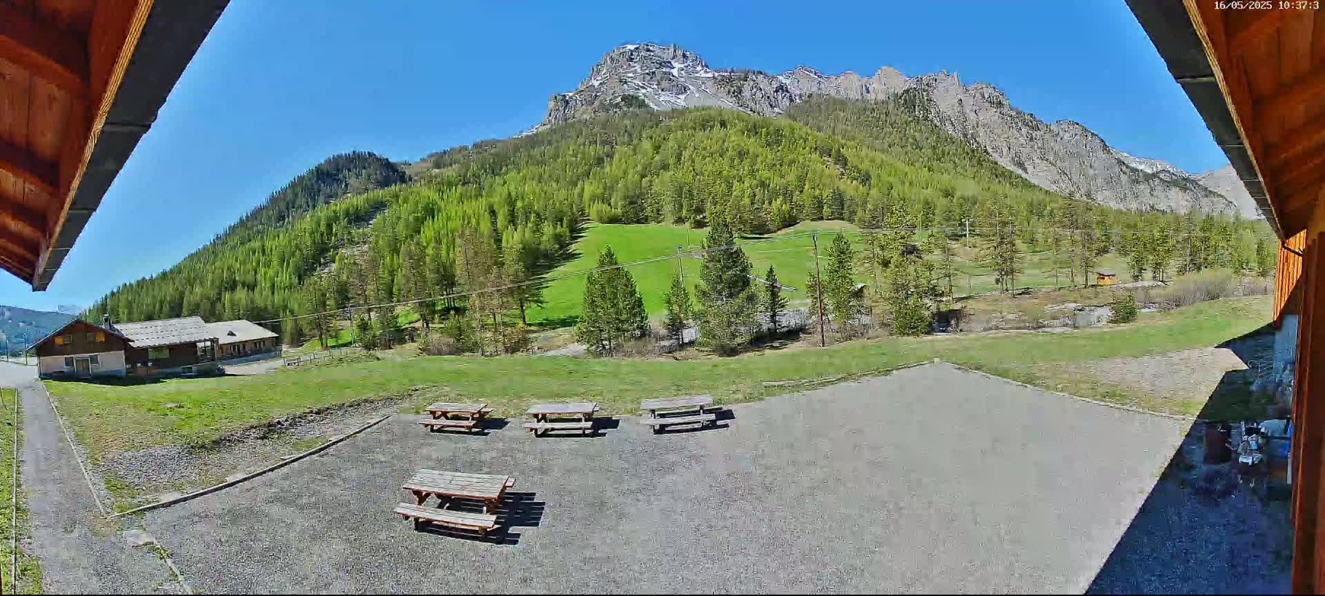 A sunny view from under a wooden overhang shows a gravel area with several picnic tables, a grassy field, a small building, and a large mountain covered with green trees in the background.