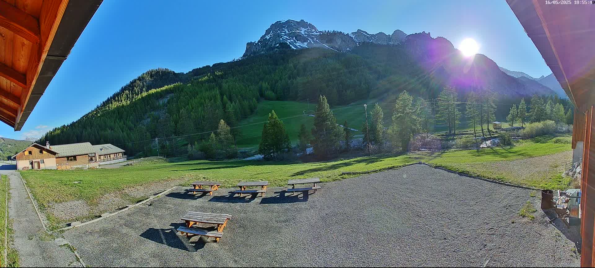 Several picnic tables sit in a gravel area overlooking a grassy field, a building, and a mountain range under a bright, sunny sky.