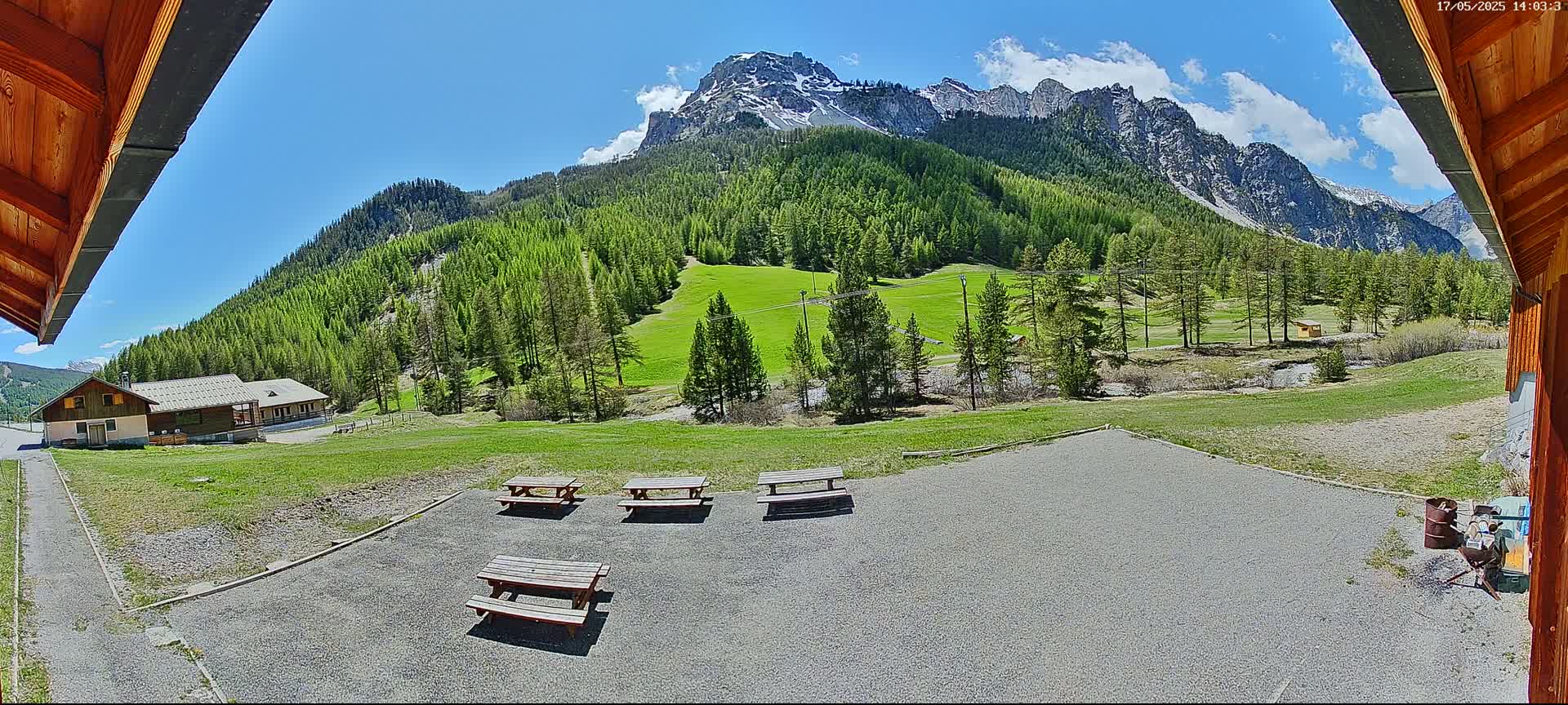 A gravel area with picnic tables overlooks a grassy field and a mountain range under a mostly sunny sky.