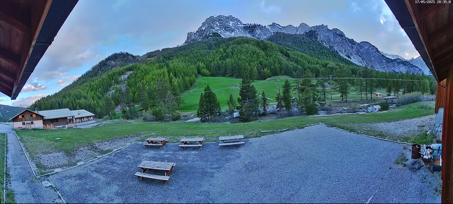 A gravel area with several picnic tables sits in front of a building, overlooking a grassy field and a mountain range with patches of snow under a partly cloudy evening sky.