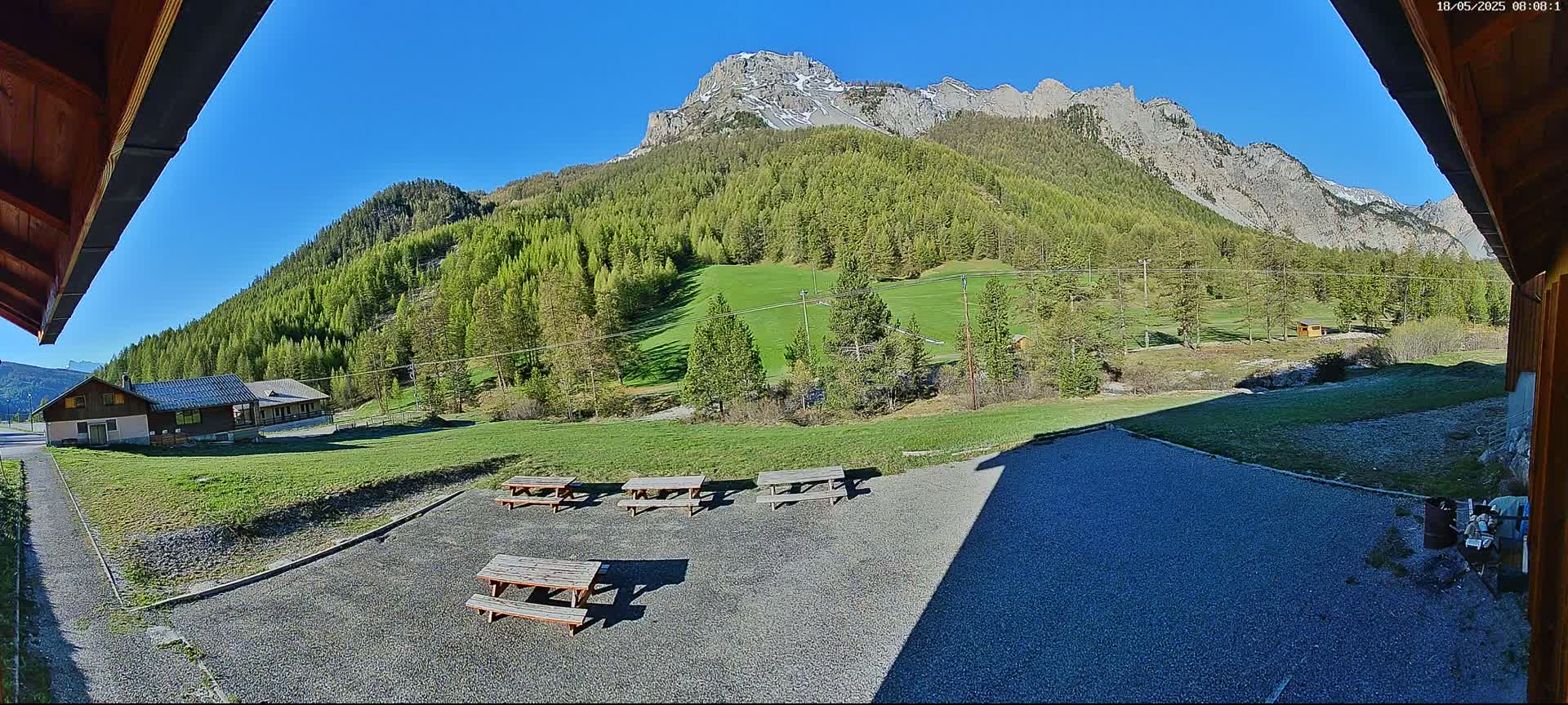 A gravel area with several picnic tables overlooks a grassy field and a mountainside covered in lush green trees under a clear blue sky.