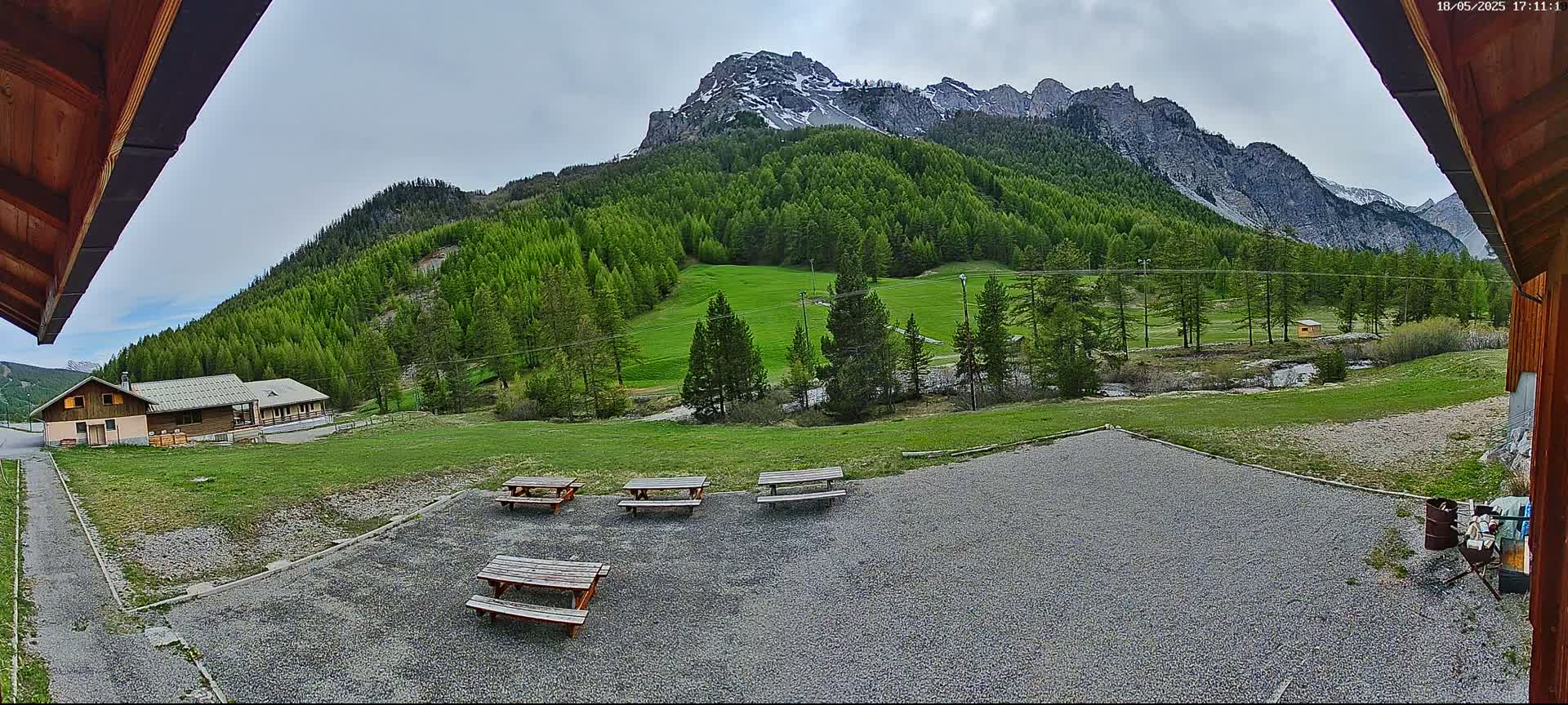 A gravel area with several picnic tables overlooks a grassy field, a small building, and a mountain range with patchy snow under a cloudy sky.