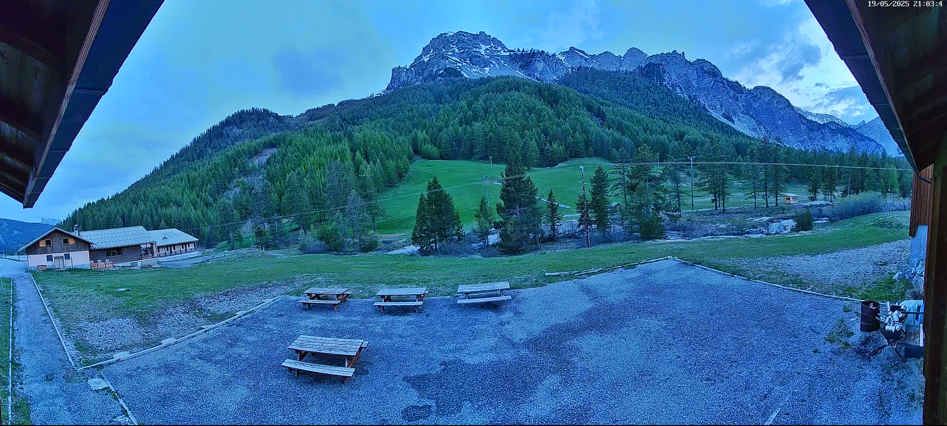 A gravel area with several picnic tables sits before a building, overlooking a grassy field and a mountain range under a twilight sky.