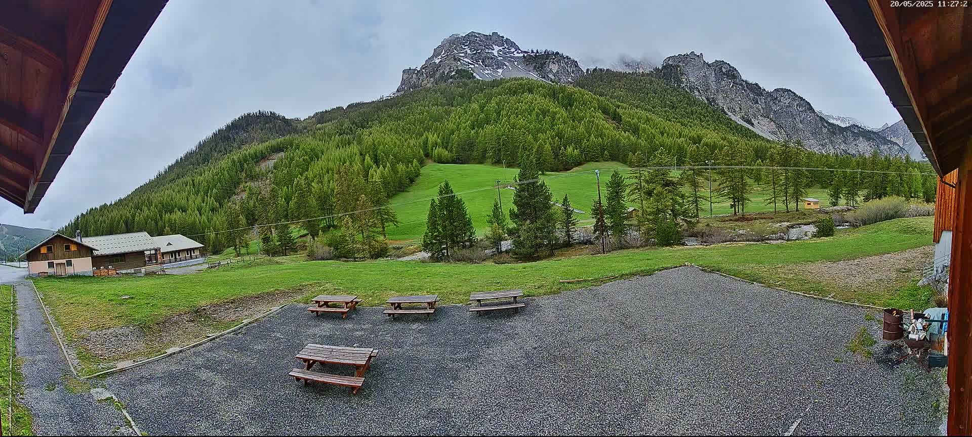Several picnic tables sit on a gravel area overlooking a grassy field, a small building, and a mountain range under a cloudy sky.