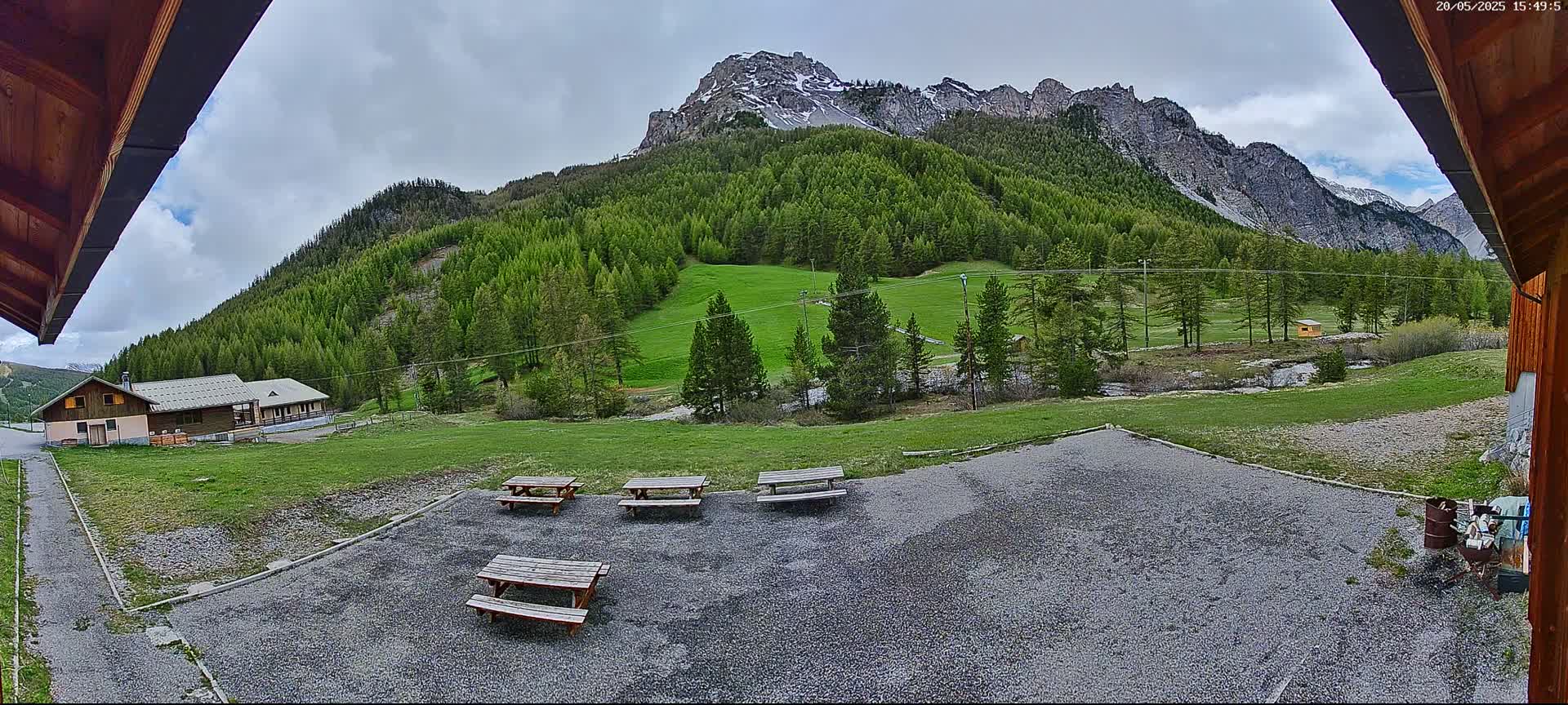 A gravel area with several picnic tables sits before a building and a verdant mountainside under a partly cloudy sky.