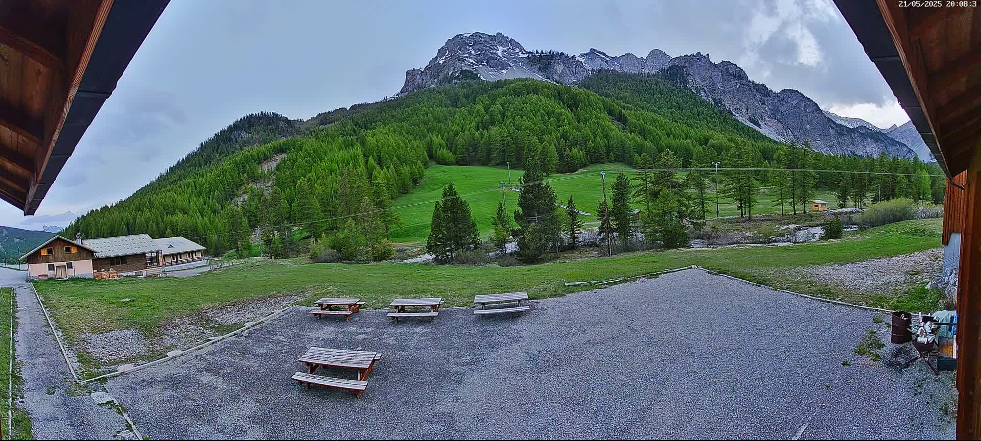 A gravel area with several picnic tables sits before a grassy field and a mountain range covered in lush green trees under a partly cloudy sky.