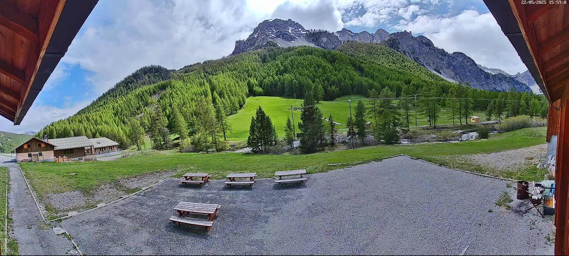 A gravel area with picnic tables overlooks a grassy valley, a small building, and a mountain range under a partly cloudy sky.
