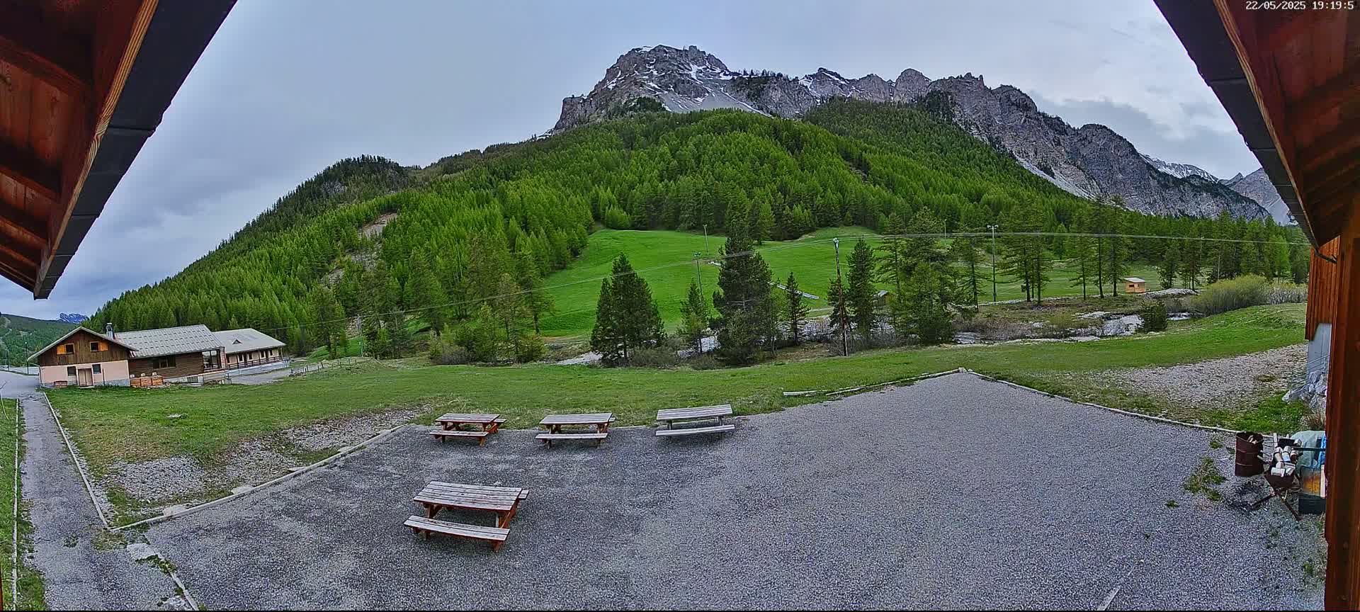 A gravel area with several picnic tables sits in front of a building, overlooking a grassy field, a forest of evergreen trees, and a mountain range under a partly cloudy sky.