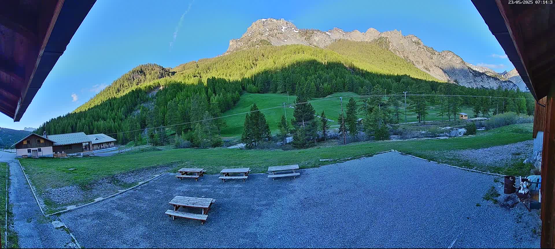 A gravel area with picnic tables is situated in front of a building, overlooking a verdant valley and a mountain range under a clear blue sky.