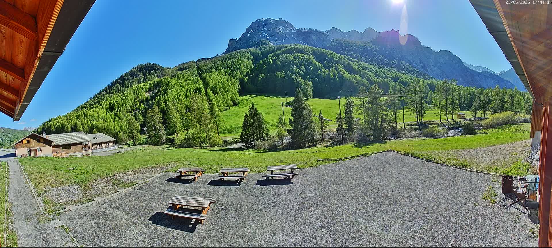 A sunny view from under a wooden awning shows a gravel area with picnic tables, a grassy field, a small building, and a mountain range in the background.
