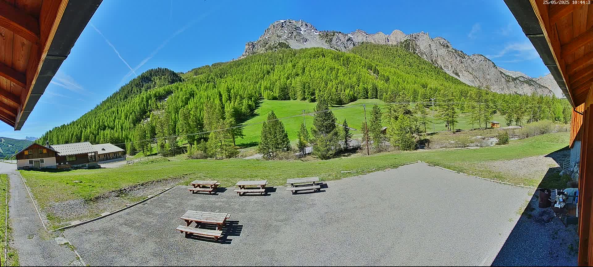 A sunny view from a patio shows several picnic tables overlooking a grassy field, a building, and a mountain range covered in lush green trees.