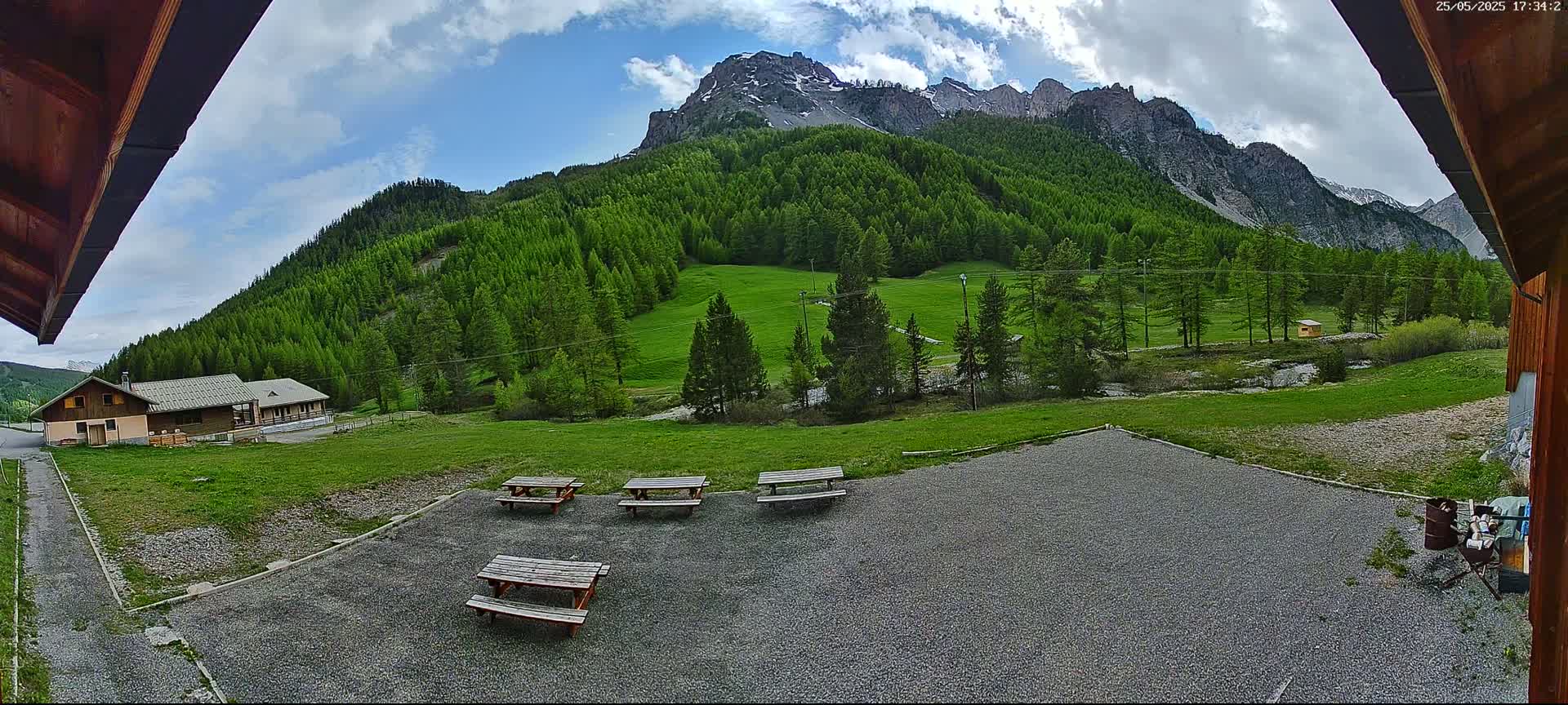 A gravel area with several picnic tables sits in front of a building, overlooking a grassy field and a mountain range covered in lush green trees under a partly cloudy sky.